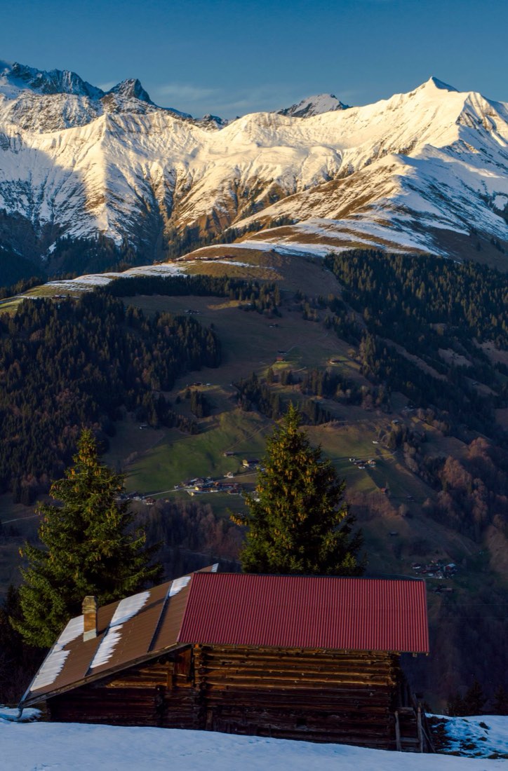 红顶原木小屋立于山坡,身后是夕照下的雪山与山谷